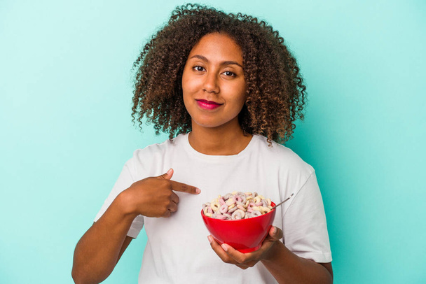Jonge Afro-Amerikaanse vrouw met een kom granen geïsoleerd op blauwe achtergrond persoon wijzend met de hand naar een shirt kopieerruimte, trots en vol vertrouwen - Foto, afbeelding