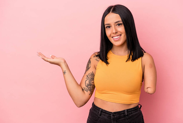 Young caucasian woman with one arm isolated on pink background showing a copy space on a palm and holding another hand on waist. - Photo, Image
