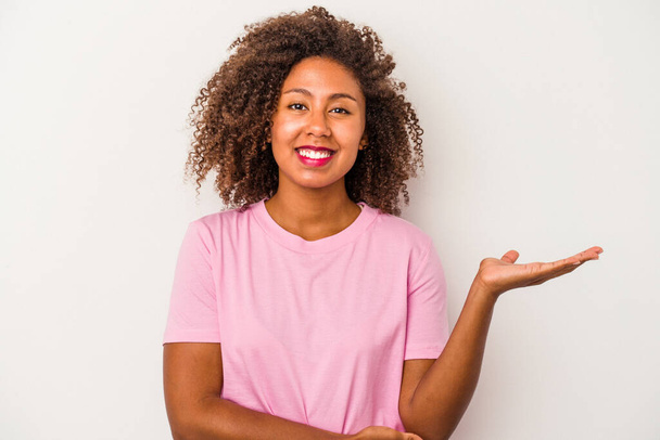 Молодая африканская американка с кудрявыми волосами на белом фоне показывает пространство для копирования на ладони и держит другую руку на талии. - Фото, изображение