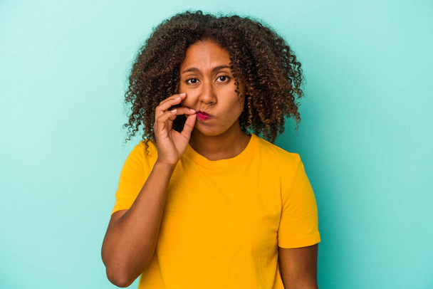 Young african american woman with curly hair isolated on blue background with fingers on lips keeping a secret. - 写真・画像