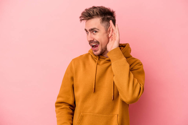 Young caucasian man isolated on pink background trying to listening a gossip. - Photo, image