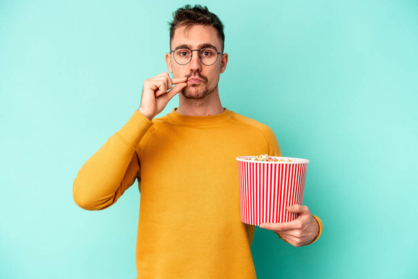 Fiatal kaukázusi férfi eszik popcorn izolált kék háttérrel ujjak az ajkakon tartja a titkot. - Fotó, kép