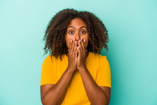 Молодая африканская американка с вьющимися волосами изолирована на синем фоне, прикрывая рот руками, выглядящими обеспокоенными.. - Фото, изображение