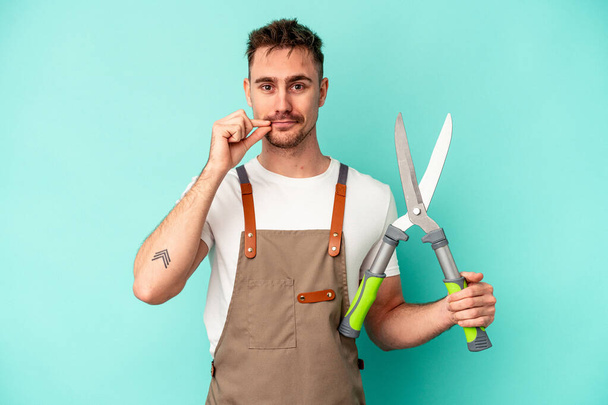 Young gardener caucasian man holding a scissors isolated on blue background with fingers on lips keeping a secret. - Fotoğraf, Görsel