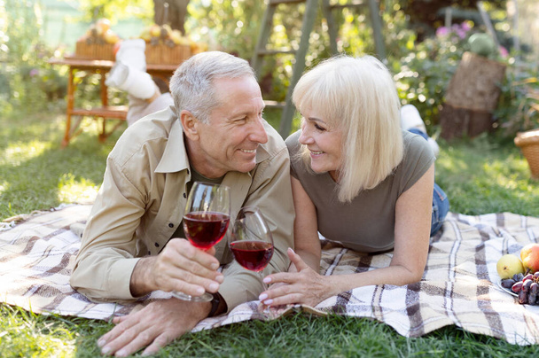 Rakastava aviopari vanhempi pari juo viiniä ulkona, makaa huopalla, kun heillä on piknik puutarhassaan - Valokuva, kuva