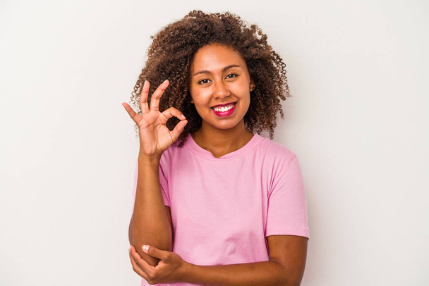 Young african american woman with curly hair isolated on white background winks an eye and holds an okay gesture with hand. - Фото, изображение