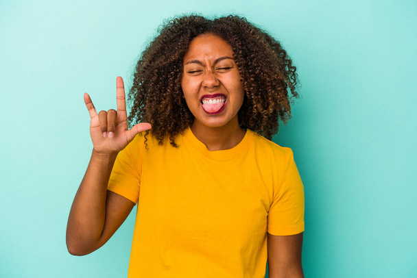 Young african american woman with curly hair isolated on blue background showing rock gesture with fingers - Foto, afbeelding