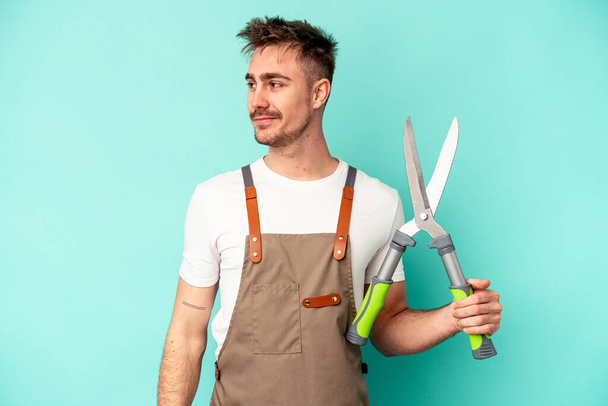 Young gardener caucasian man holding a scissors isolated on blue background looks aside smiling, cheerful and pleasant. - Foto, afbeelding
