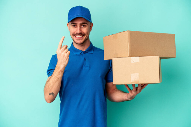 Young delivery caucasian man isolated on blue background pointing with finger at you as if inviting come closer. - Photo, Image