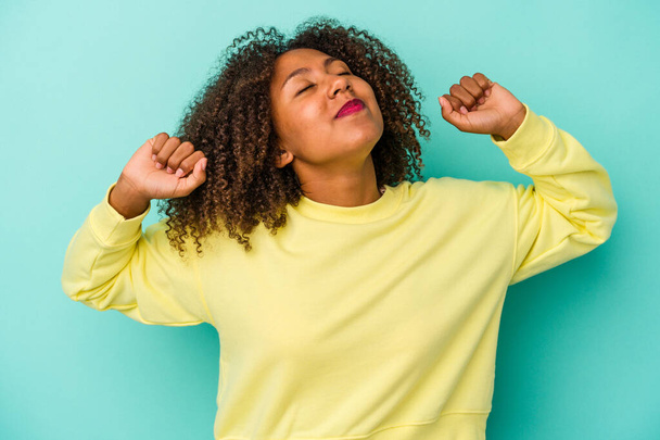 Young african american woman with curly hair isolated on blue background stretching arms, relaxed position. - Photo, Image