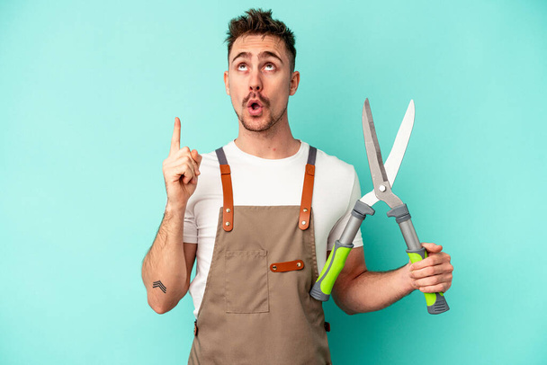 Young gardener caucasian man holding a scissors isolated on blue background pointing upside with opened mouth. - Foto, immagini