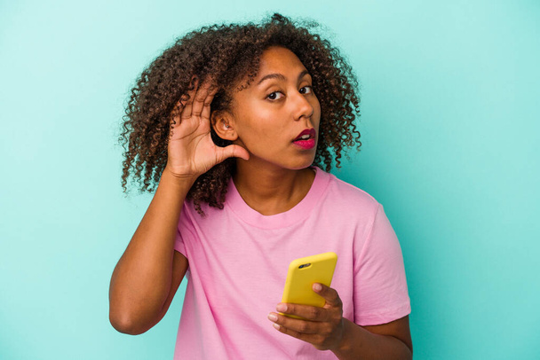 Mujer afroamericana joven sosteniendo un teléfono móvil aislado sobre fondo azul tratando de escuchar un chisme. - Foto, Imagen
