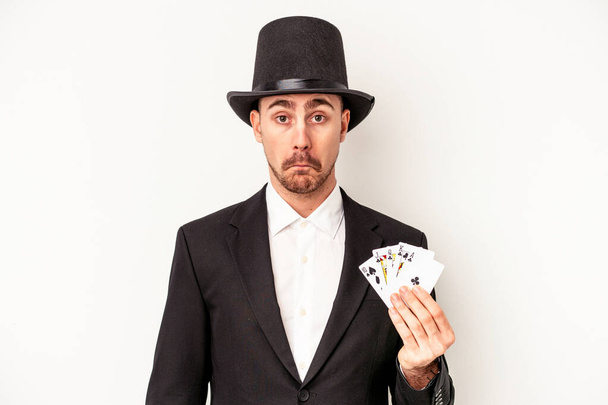 Mladý běloch čaroděj muž drží magické karty izolované na bílém pozadí pokrčí rameny a otevřené oči zmatené. - Fotografie, Obrázek