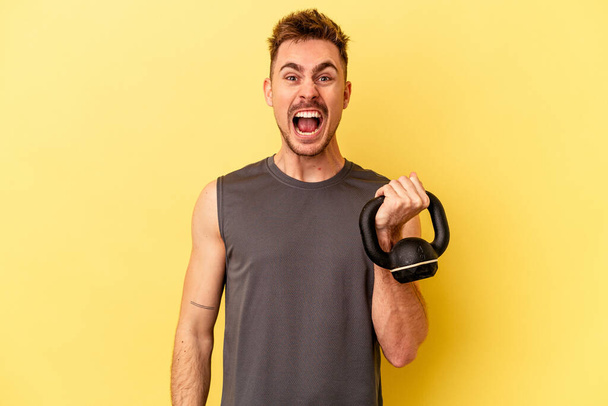 Young sport man holding a dumbbell isolated on yellow background screaming very angry and aggressive. - Photo, Image