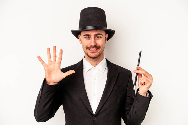 Giovane mago caucasico uomo tenendo una bacchetta isolata su sfondo bianco sorridente allegro mostrando il numero cinque con le dita. - Foto, immagini
