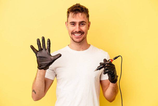 Young caucasian tattoo maker man isolated on yellow background smiling cheerful showing number five with fingers. - Photo, Image