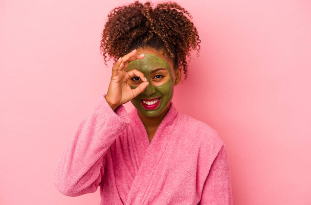 Mujer afroamericana joven con una bata de baño y mascarilla facial aislado sobre fondo rosa emocionado mantener el gesto de ok en el ojo. - Foto, imagen