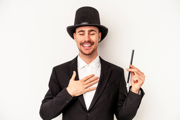 Young caucasian wizard man holding a wand isolated on white background laughs out loudly keeping hand on chest. - Photo, Image