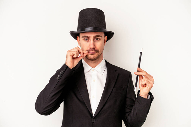 Jeune magicien caucasien tenant une baguette isolée sur fond blanc avec les doigts sur les lèvres gardant un secret. - Photo, image