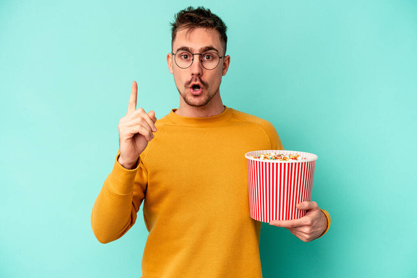 Young caucasian man eating popcorn isolated on blue background having an idea, inspiration concept. - Foto, Bild