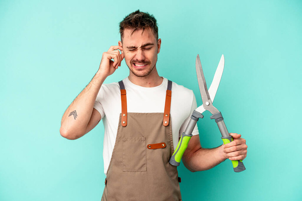 Молодий садівник білий чоловік тримає ножиці ізольовані на синьому фоні, покриваючи вуха руками
. - Фото, зображення