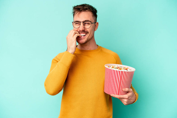 Jonge blanke man eet popcorn geïsoleerd op blauwe achtergrond bijtende vingernagels, nerveus en zeer angstig. - Foto, afbeelding