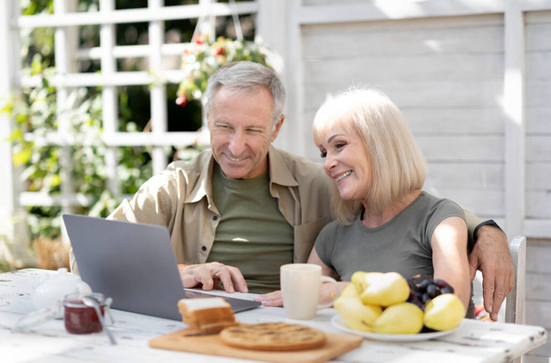 ラップトップを屋外で使用して幸せな高齢者のカップル,自宅のテラスに座って、インターネットサーフィンやビデオ通話を持っている - 写真・画像