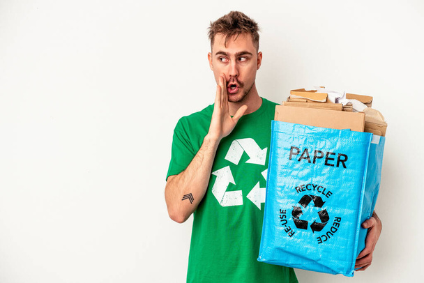 Junge kaukasische Mann Recyclingpapier isoliert auf weißem Hintergrund sagt eine geheime heiße Bremse Nachrichten und Blick zur Seite - Foto, Bild