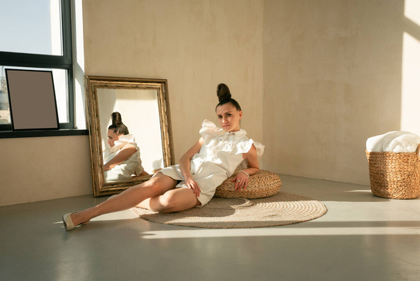 Tyttö valkoinen mekko beige sisustus moderni kampausNuori kaunis ukrainalainen nainen istuu lattialla pyöreä matto näyttää peiliin hiukset kerätty tyylikäs kampaus on ruskeat silmät kauniit kasvot hänen käsivartensa rannerengas - Valokuva, kuva