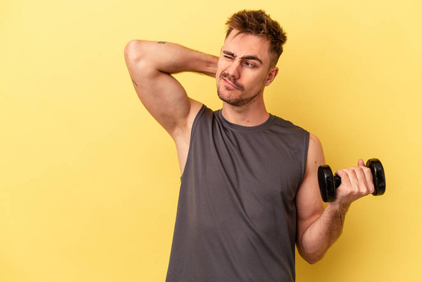 Young sport man holding a dumbbell isolated on yellow background touching back of head, thinking and making a choice. - Photo, Image
