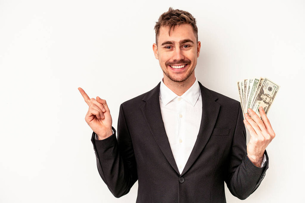 Jeune homme d'affaires caucasien tenant des billets de banque isolés sur fond blanc souriant et pointant du doigt, montrant quelque chose à blanc. - Photo, image
