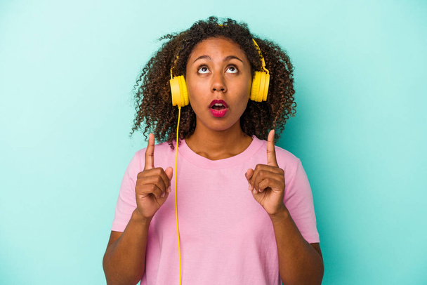 Young african american woman listening to music isolated on blue background pointing upside with opened mouth. - Foto, Imagen