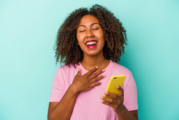 Young african american woman holding a mobile phone isolated on blue background laughs out loudly keeping hand on chest. - Photo, Image