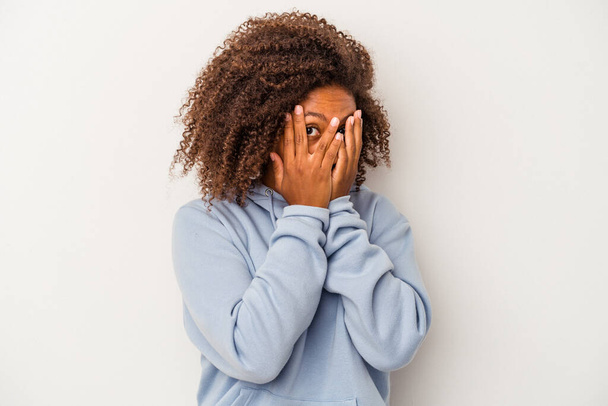 Jeune femme afro-américaine aux cheveux bouclés isolés sur fond blanc clignent des yeux à travers les doigts effrayés et nerveux. - Photo, image