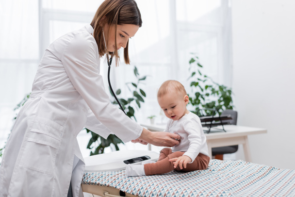 Junger Arzt untersucht Baby mit Stethoskop auf Medizinercouch in Klinik   - Foto, Bild