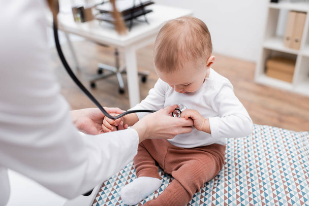 Размытый врач осматривает ребенка со стетоскопом на медицинском диване в клинике  - Фото, изображение
