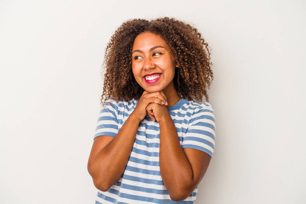 jonge afrikaanse amerikaanse vrouw met krullend haar geïsoleerd op witte achtergrond houdt handen onder kin, is op zoek gelukkig opzij. - Foto, afbeelding