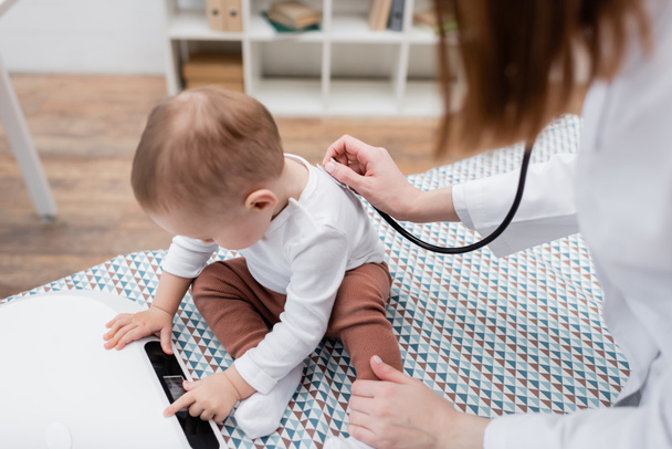 Hausarzt untersucht kleinen Jungen mit Stethoskop nahe elektronischer Waage in Klinik  - Foto, Bild