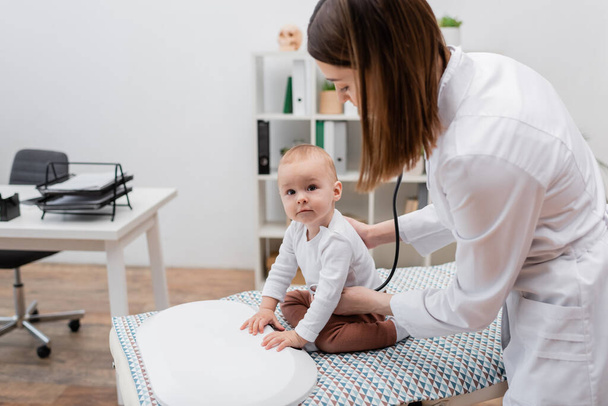 Nuori perhelääkäri tutkii rintakehän vauva poika lähellä sähköisen mittakaavan lääketieteen sohvalla  - Valokuva, kuva
