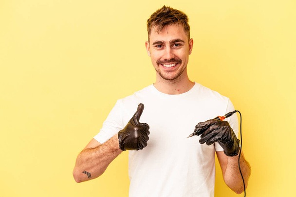 Молодой белый татуировщик мужчина изолирован на желтом фоне улыбаясь и поднимая большой палец вверх - Фото, изображение