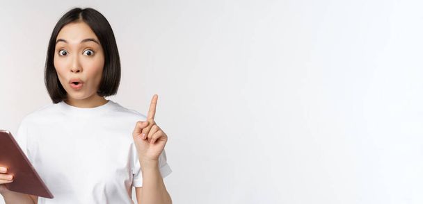 Enthusiastic asian woman with tablet, raising finger and looking amazed, pointing up, standing over white background - Foto, Bild
