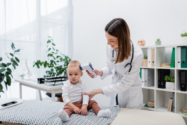 Medico di famiglia positivo che controlla la temperatura del bambino con pirometro in clinica  - Foto, immagini