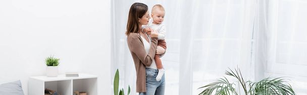 Young woman holding baby son near plant at home, banner  - Photo, Image