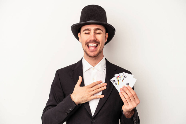 Giovane mago caucasico uomo in possesso di una carta magica isolato su sfondo bianco ride ad alta voce mantenendo la mano sul petto. - Foto, immagini