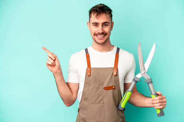 Young gardener caucasian man holding a scissors isolated on blue background smiling and pointing aside, showing something at blank space. - Photo, Image