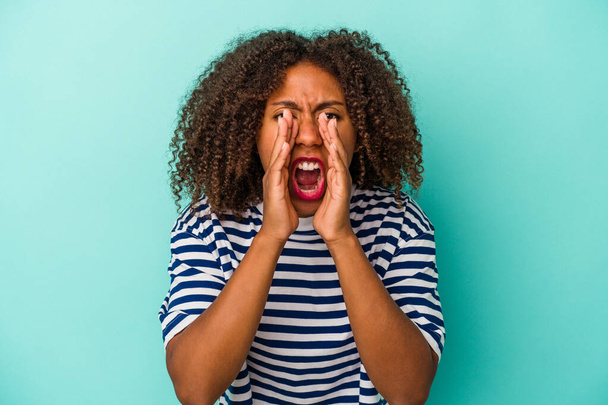 Young african american woman with curly hair isolated on blue background shouting excited to front. - 写真・画像