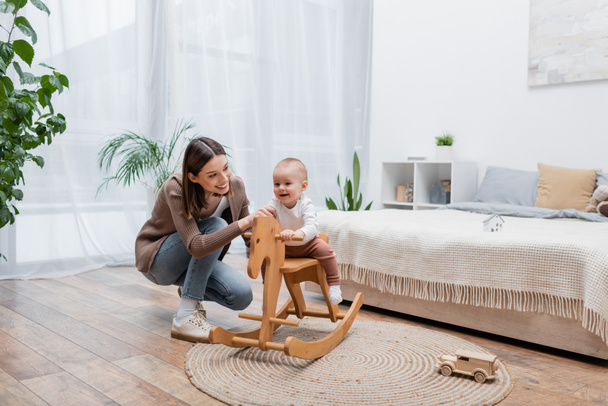 Positive baby boy sitting on rocking horse near mom in bedroom  - Photo, Image