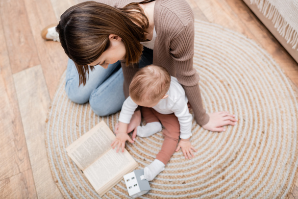 Nad hlavou pohled na ženu čtení knihy v blízkosti rozmazané dítě syna na podlaze doma  - Fotografie, Obrázek