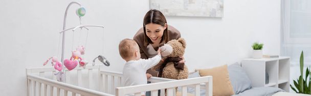 Young mother holding soft toy near baby boy in crib, banner  - Photo, Image