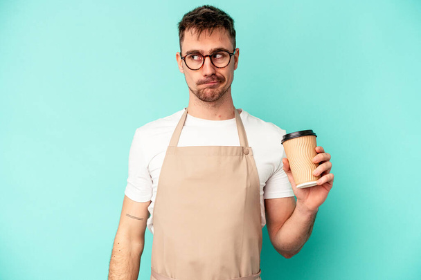Young store clerk man holding a coffee isolated on blue background confused, feels doubtful and unsure. - 写真・画像